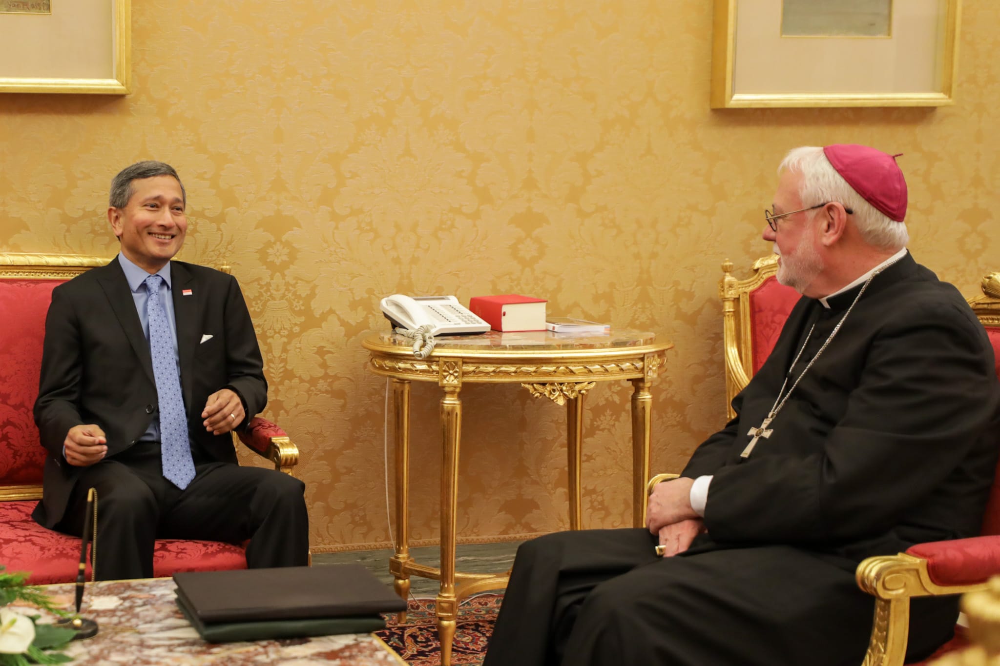 Man in suit facing Bishop in formal black robe with cross pendant in ornate room.